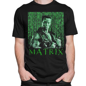 Puede incluir: Camiseta negra con un gráfico verde de un hombre con un fondo temático de Matrix. La palabra "MATRIX" está impresa en verde debajo de la imagen. La camiseta es de un material suave.
