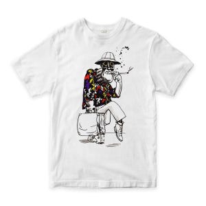 May include: White t-shirt with a black and white illustration of a seated figure wearing a hat and smoking. The figure is dressed in a colourful floral shirt, white trousers, and boots, holding a suitcase. Black ink splatters are above the figure.