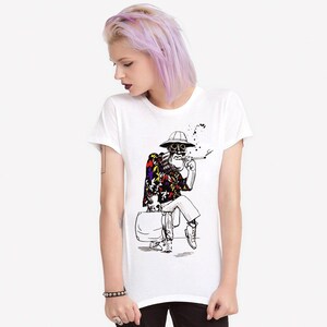 May include: White crew neck t-shirt with a black and white illustration of a skeleton figure. The figure is wearing a patterned shirt, hat, and boots, smoking a cigarette and sitting on a suitcase.