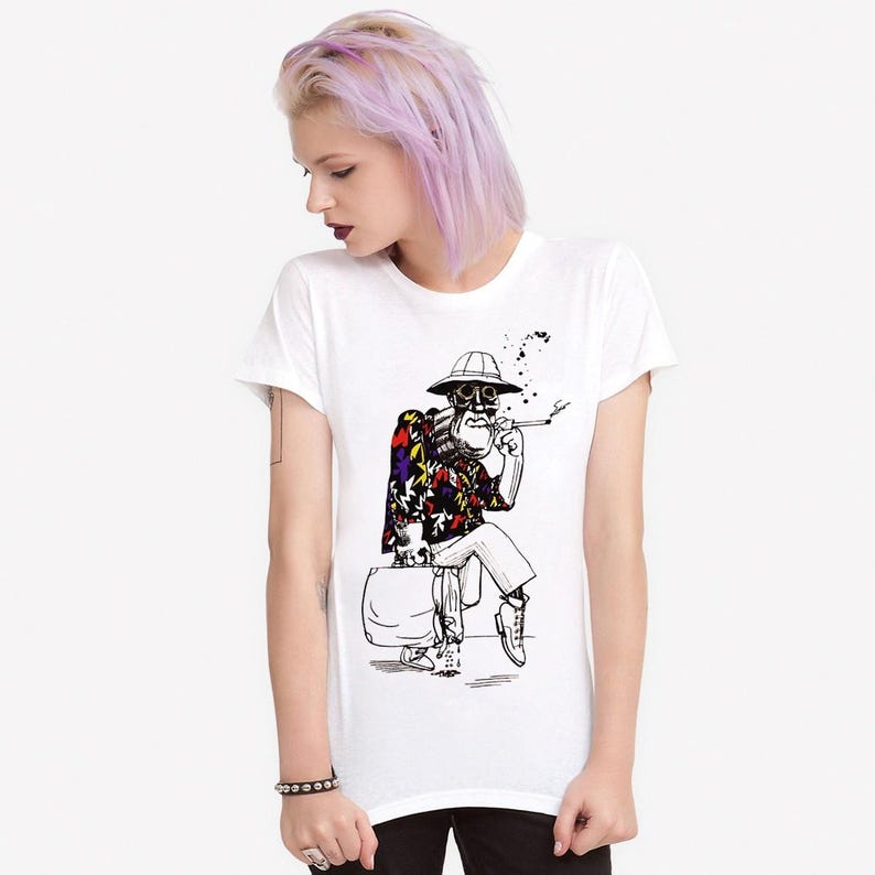 May include: White crew neck t-shirt with a black and white illustration of a skeleton figure. The figure is wearing a patterned shirt, hat, and boots, smoking a cigarette and sitting on a suitcase.