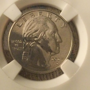 Może przedstawiać: Srebrna amerykańska moneta o wartości ćwierć dolara z napisem "LIBERTY" i "IN GOD WE TRUST" na awersie. Na monecie widnieje profil George'a Washingtona i data "2022".