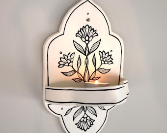 Può includere: Un'applique da parete in ceramica bianca con un design floreale nero. L'applique ha un ripiano curvo per contenere una candela, e la candela è accesa, proiettando un bagliore caldo. Il design è simmetrico e presenta fiori stilizzati.
