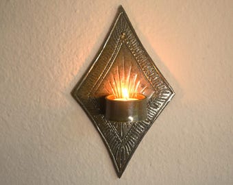 May include: A diamond-shaped wall sconce made of dark metal with a textured, sunburst-like pattern radiating from the center. A small, cylindrical candle holder is attached to the front, containing a lit tea light candle with a warm orange glow.