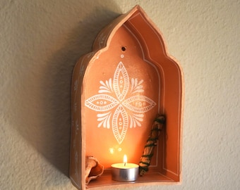 May include: A terracotta-colored wall-mounted shelf with an arched top, featuring white hand-painted floral patterns. Inside the shelf sits a small lit tea light candle, a seashell, and a bundle of dried herbs tied with green string.