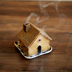 May include: A small, tan ceramic house-shaped incense burner with a textured roof and a chimney. Wisps of white smoke rise from the chimney and the arched doorway. The structure sits on a small, square base with a black and white patterned edge.