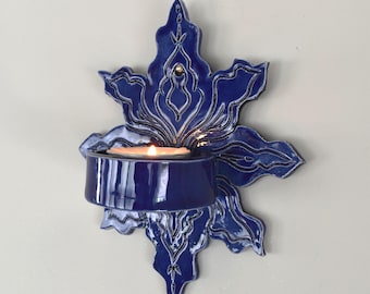 Può includere: Un'applique da parete in ceramica smaltata blu profondo caratterizzata da una silhouette floreale simmetrica a forma di stella, con un supporto circolare centrale contenente una candela tea light accesa. La finitura lucida esalta i motivi.