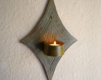 May include: A diamond-shaped wall sconce featuring a grey, textured metal backplate with etched sunburst patterns and a small hole at the top. A cylindrical, dark-toned metal cup is attached to the center, holding a small, lit tea light candle.