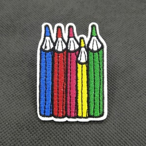 Puede incluir: Parche bordado con cinco lápices de colores en azul, rojo, rosa, amarillo y verde. Cada lápiz tiene una punta blanca y está delineado en negro. El parche tiene un borde blanco sobre un fondo gris oscuro.