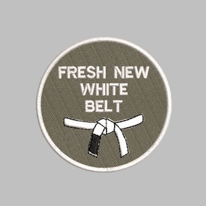 Op de afbeelding: Geborduurde patch met de tekst "FRESH NEW WHITE BELT" in witte letters. De patch heeft een wit en zwart karate-riemontwerp op een cirkelvormige, olijfgroene achtergrond met een witte rand. Voor liefhebbers van vechtsporten.