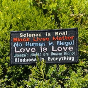 May include: Black embroidered patch with the text "Science is Real, Black Lives Matter, No Human is Illegal, Love is Love, Women's Rights are Human Rights, Kindness is Everything".