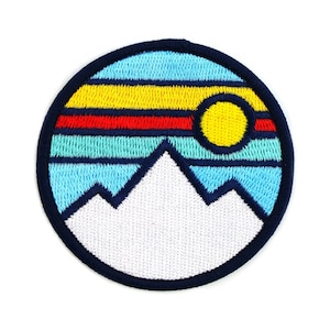 May include: A round embroidered patch with a white mountain range, a yellow sun, and a blue sky with red and yellow stripes.