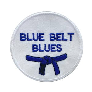 May include: A round, white patch with the words "BLUE BELT BLUES" embroidered in blue. A blue belt graphic is embroidered below the text. The patch has a white stitched border.