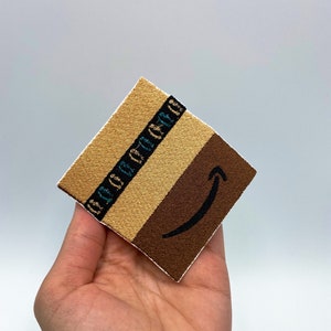 Puede incluir: Un parche de tela marrón y beige con una franja azul y blanca y un logotipo de Amazon marrón.