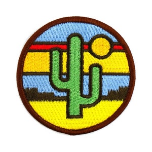 May include: Embroidered patch with a green cactus in a desert scene, a yellow sun, and a blue sky. The design has a brown border and a red stripe, suggesting a Southwestern theme.