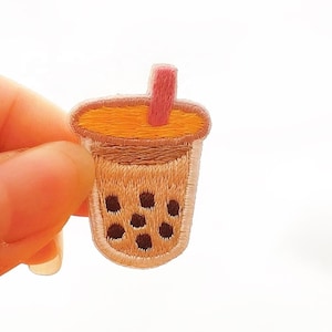 May include: Embroidered patch featuring a bubble tea drink. The patch has a light brown cup with dark brown boba pearls, an orange-yellow top, and a pink straw. The patch is held by a hand.