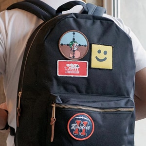 Peut inclure: Sac à dos noir orné de plusieurs écussons. Les écussons comprennent un smiley, un personnage de dessin animé dans un viseur, un rectangle rouge avec un éléphant et les mots "NEVER FORGET", et un écusson en forme de ballon de football avec le mot "Raiders".