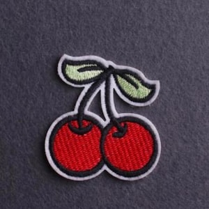 Peut inclure: Un écusson brodé représentant deux cerises rouges rondes avec des contours noirs et deux feuilles vert clair. Le motif est bordé d'un épais contour blanc, créant un contraste marqué sur la surface de fond texturée gris foncé.