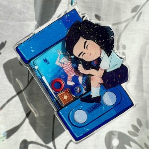May include: A blue cassette tape-shaped keychain with a cartoon illustration of a girl sleeping. The keychain has a blue star-shaped clasp. The illustration includes a figure in a striped shirt and a fish.