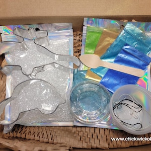 May include: A craft kit with two metal cookie cutters shaped like dolphins, a bag of clear glitter, four small bags of colored glitter, a wooden spoon, a clear plastic container with blue glitter, and a clear plastic container with a string.