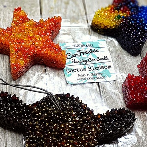 May include: Three star-shaped car air fresheners in orange, black, and red. The orange air freshener is labeled "Car Freshie Hanging Car Candle Cactus Blossom Made in Oklahoma www.chickwickokc.com"
