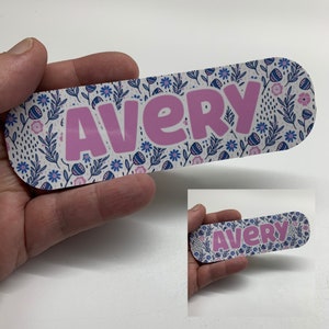 May include: A personalised nameplate with the name "Avery" in pink bubble letters. The background features a floral pattern with blue and pink flowers and green leaves on a white background. The nameplate is held in a hand.