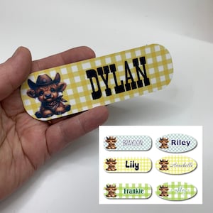May include: A set of six personalized iron-on labels featuring a cartoon cow wearing a cowboy hat. The labels are in various colors and feature the names Braydon, Riley, Lily, Annabelle, Frankie, and Ashley.