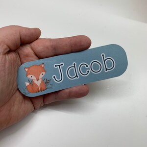 May include: A blue name tag with rounded edges featuring the name "Jacob" in white outlined letters. A cartoon fox with orange fur and a white belly is on the left side of the tag, with green leaves.