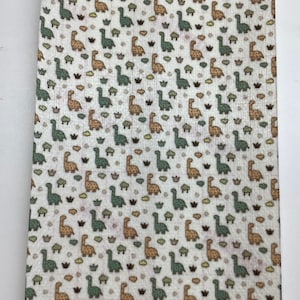 May include: A rectangular piece of fabric featuring a repeating pattern of cartoon dinosaurs in green and tan on a cream background. Small brown and yellow cloud and bush-like shapes are scattered throughout the design.
