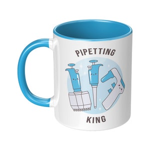 May include: White ceramic mug with a blue rim and handle. The mug has a graphic of three pipettes with faces and the text "PIPETTING KING".