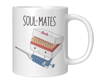 Puede incluir: Taza de cerámica blanca con una pipeta azul y blanca y una caja de puntas de pipeta de dibujos animados con un lazo rosa. El texto "SOULMATES" está impreso en la taza.