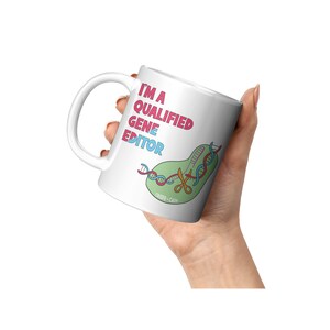 Peut inclure: Une tasse en c&eacute;ramique blanche avec une illustration rose, bleue et verte d'un brin d'ADN coup&eacute; par des ciseaux. Le texte "I'm a Qualified Gene Editor" est &eacute;crit en rose et bleu sur la tasse.