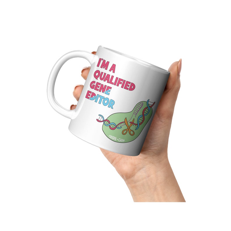 Peut inclure: Une tasse en c&eacute;ramique blanche avec une illustration rose, bleue et verte d'un brin d'ADN coup&eacute; par des ciseaux. Le texte "I'm a Qualified Gene Editor" est &eacute;crit en rose et bleu sur la tasse.