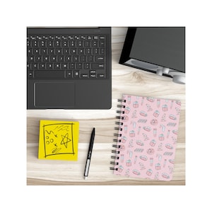Peut inclure: Un carnet &agrave; spirales rose avec un motif sur le th&egrave;me des sciences. Le motif comprend des tubes &agrave; essai, des b&eacute;chers, des microscopes et d'autres &eacute;quipements scientifiques. Le carnet est sur une surface en bois avec un post-it jaune et un stylo noir.