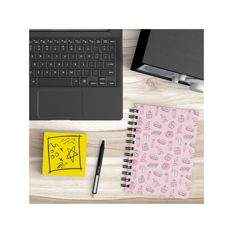 Peut inclure: Un carnet &agrave; spirales rose avec un motif sur le th&egrave;me des sciences. Le motif comprend des tubes &agrave; essai, des b&eacute;chers, des microscopes et d'autres &eacute;quipements scientifiques. Le carnet est sur une surface en bois avec un post-it jaune et un stylo noir.