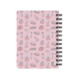 Peut inclure: Un carnet rose avec une reliure spirale. La couverture pr&eacute;sente un motif r&eacute;p&eacute;t&eacute; d'illustrations sur le th&egrave;me des sciences, notamment des b&eacute;chers, des tubes &agrave; essai, des microscopes et des bo&icirc;tes de P&eacute;tri.