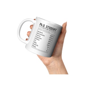 May include: White ceramic mug with a black graphic of a nutrition facts label for a Ph.D. student. The label lists the percentage of various items such as hard work, determination, passion, exhaustion, caffeine, cry, sleep, social life, and money.