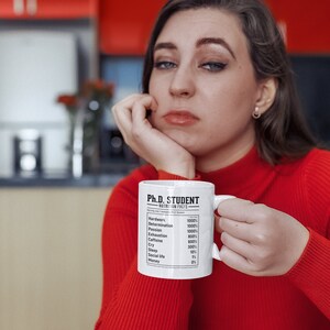 May include: White ceramic mug with a black and white graphic that reads "Ph.D. Student Nutrition Facts" with a list of items and percentages. The mug is held by a person wearing a red sweater.