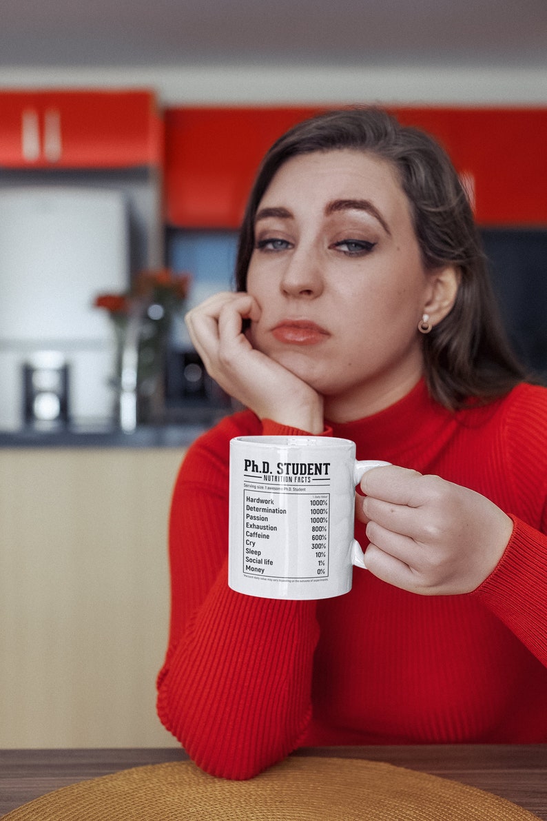 May include: White ceramic mug with a black and white graphic that reads "Ph.D. Student Nutrition Facts" with a list of items and percentages. The mug is held by a person wearing a red sweater.