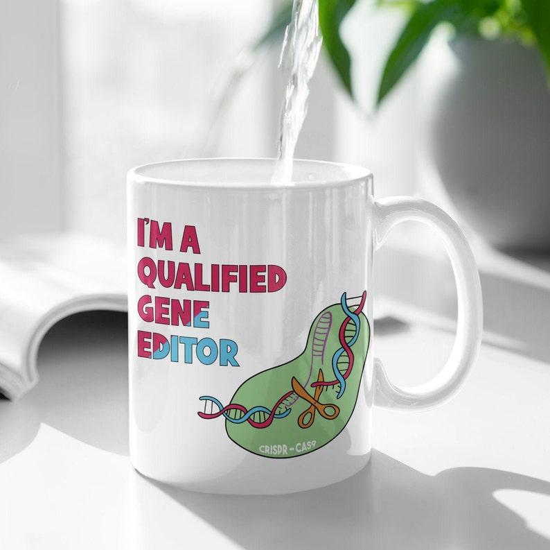 Peut inclure: Une tasse en c&eacute;ramique blanche avec une illustration verte, bleue et rouge d'une cha&icirc;ne d'ADN coup&eacute;e par des ciseaux. Le texte "I'm a Qualified Gene Editor" est imprim&eacute; en rose et rouge sur la tasse. Le texte "CRISPR-Cas9" est imprim&eacute; en noir au bas de l'illustration.
