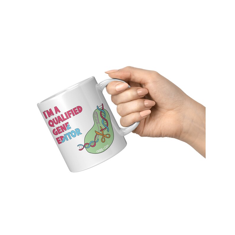 Peut inclure: Une tasse en c&eacute;ramique blanche avec une illustration rose, bleue et verte d'une cha&icirc;ne d'ADN coup&eacute;e par des ciseaux. Le texte "I'm a Qualified Gene Editor" est imprim&eacute; en rose et noir sur la tasse.