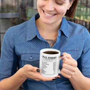 May include: White ceramic mug with a black and white graphic that reads "Ph.D. Student Nutrition Facts" and lists various items with percentages. The mug is being held by a person wearing a blue denim shirt.