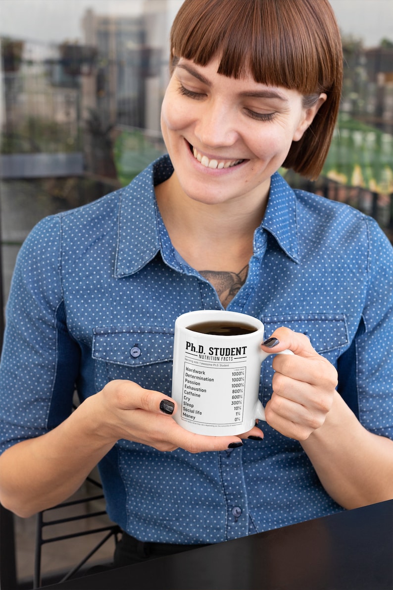 May include: White ceramic mug with a black and white graphic that reads "Ph.D. Student Nutrition Facts" and lists various items with percentages. The mug is being held by a person wearing a blue denim shirt.