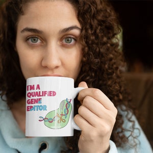 Peut inclure: Une tasse en c&eacute;ramique blanche avec une illustration verte, rose et bleue d'une cha&icirc;ne d'ADN coup&eacute;e avec des ciseaux. Le texte "I'm a Qualified Gene Editor" est imprim&eacute; en rose et noir sur la tasse.