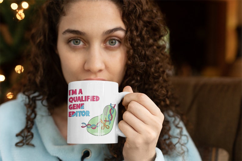 Peut inclure: Une tasse en c&eacute;ramique blanche avec une illustration verte, rose et bleue d'une cha&icirc;ne d'ADN coup&eacute;e avec des ciseaux. Le texte "I'm a Qualified Gene Editor" est imprim&eacute; en rose et noir sur la tasse.