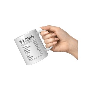 May include: White ceramic mug with a black and white graphic of a nutrition facts label for a Ph.D. student. The label lists the percentage of different elements needed for a Ph.D. student, such as hard work, determination, passion, exhaustion, caffeine, cry, sleep, social life, and money.