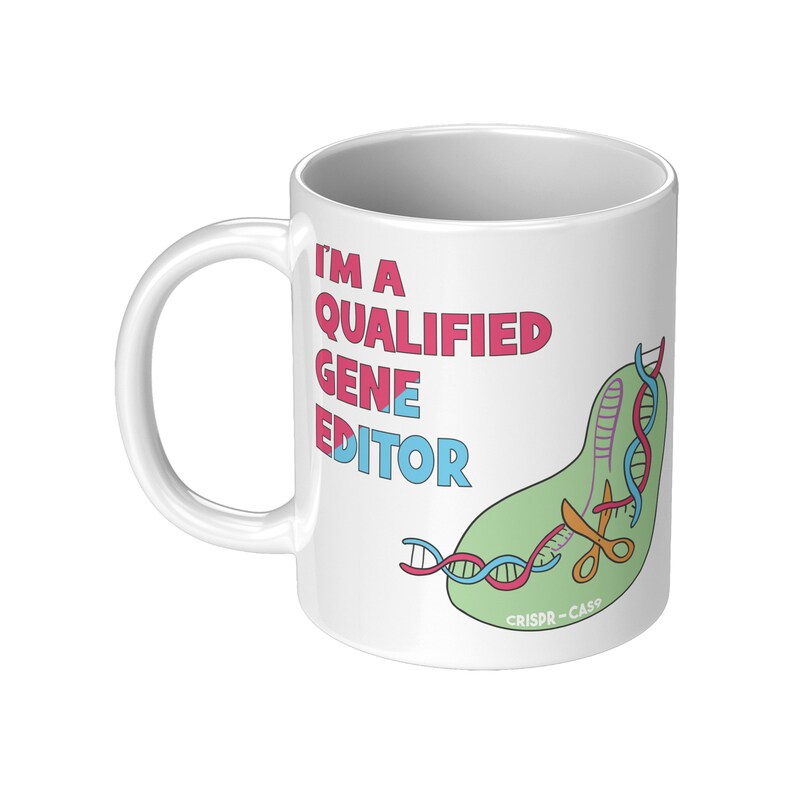 Peut inclure: Une tasse en c&eacute;ramique blanche avec une illustration verte, bleue et rouge d'une cha&icirc;ne d'ADN coup&eacute;e par des ciseaux. Le texte "I'm a Qualified Gene Editor" est imprim&eacute; en rose et bleu sur la tasse. Le texte "CRISPR-Cas9" est imprim&eacute; en noir au bas de l'illustration.