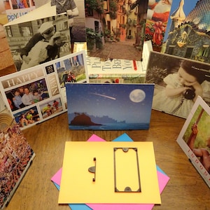 May include: A collection of greeting cards displayed on a wooden table. The cards feature a variety of images, including a family portrait, a night sky with a moon and a shooting star, a cityscape, and a child on a phone. The cards are arranged in a semi-circle around a yellow card with a black frame.
