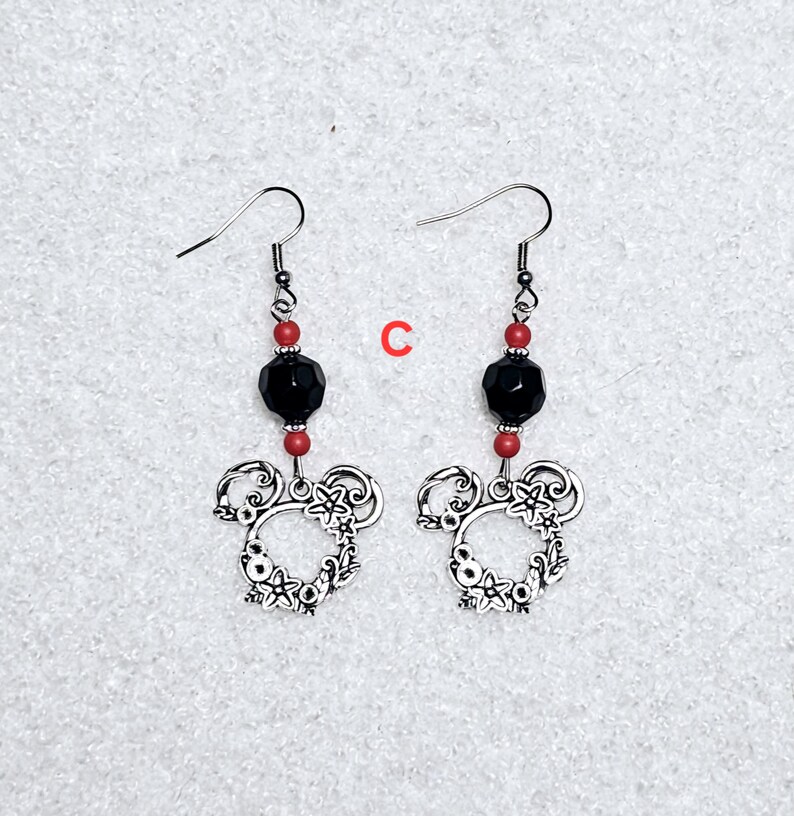 K&ouml;nnte beinhalten: Ein Paar silberne Ohrringe mit einer schwarzen facettierten Perle und roten Perlen. Die Ohrringe haben ein silbernes Mickey-Maus-Kopfdesign.