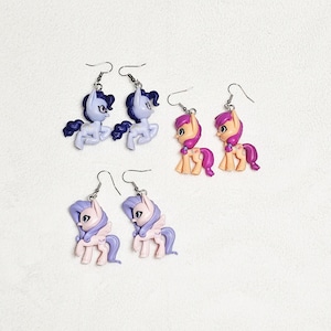 May include: Three pairs of dangle earrings featuring miniature pony figurines. One pair is light purple, one pair is peach with magenta manes, and one pair is light pink with purple manes. Each earring has a silver-toned metal hook for attachment.