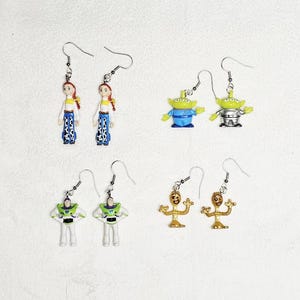 May include: A collection of novelty earrings featuring characters from the animated film Toy Story. The earrings include Jessie, the aliens, Buzz Lightyear, and Forky. Each earring has a silver hook.